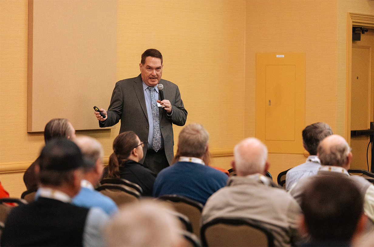 A man holding a microphone presenting to a room full of people.